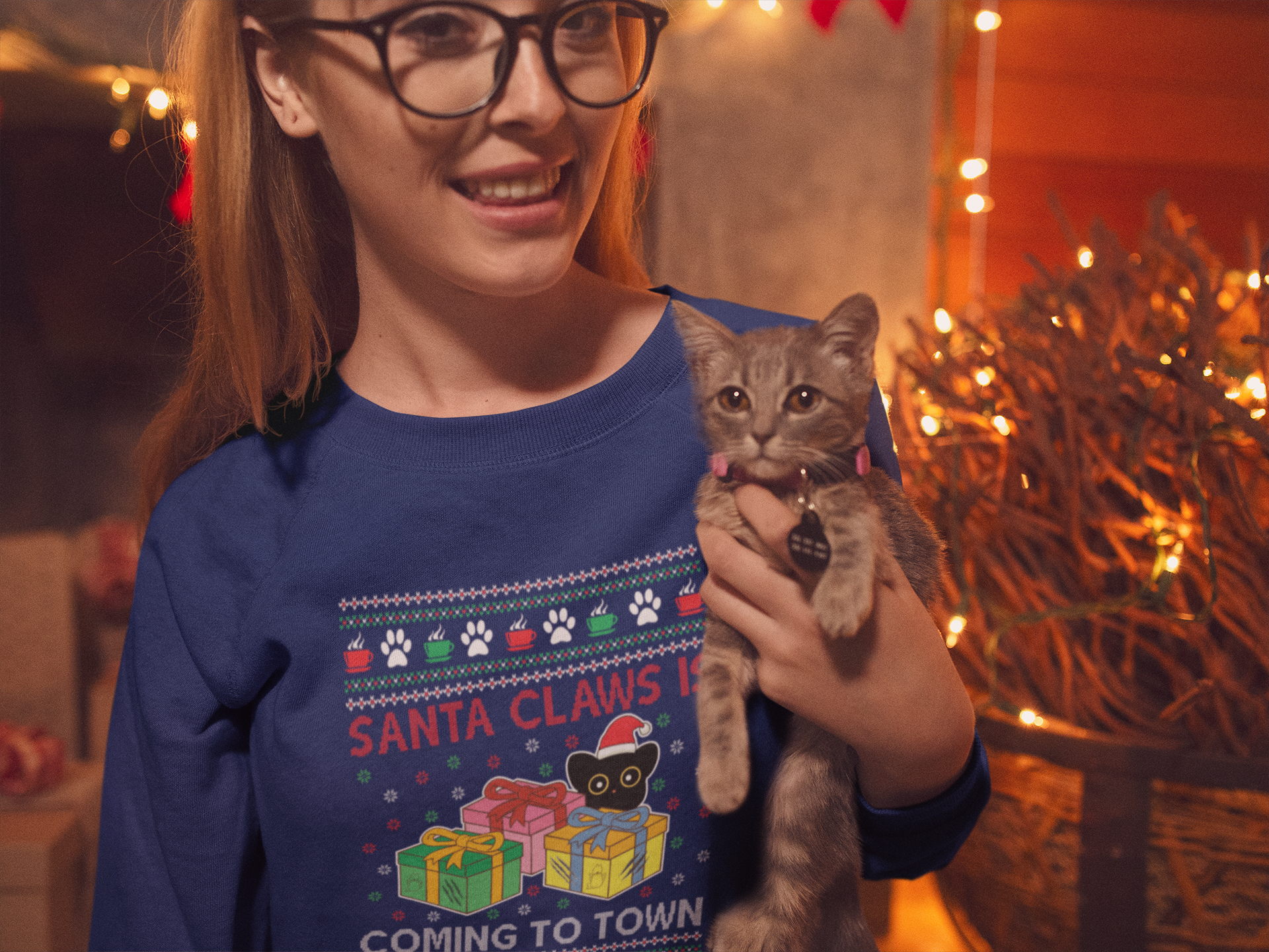 Woman wearing Purrcolate's ugly pawliday sweatshirt in the color Silent Night. She is holding a cat with Christmas decorations in the background