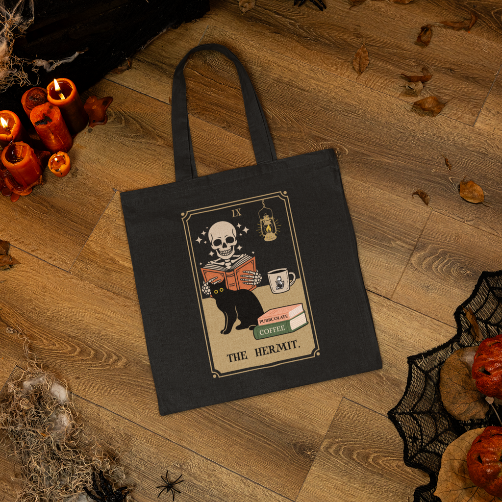 This tote features a parody of the tarot card The Hermit as a skeleton reading a book with a cat and a cup of coffee. In this image the tote is lying on the floor of a haunted house.