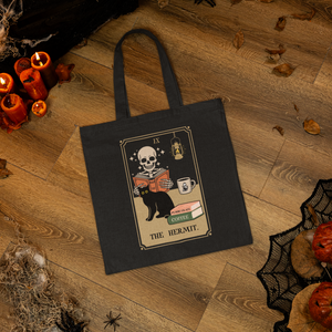 This tote features a parody of the tarot card The Hermit as a skeleton reading a book with a cat and a cup of coffee. In this image the tote is lying on the floor of a haunted house.