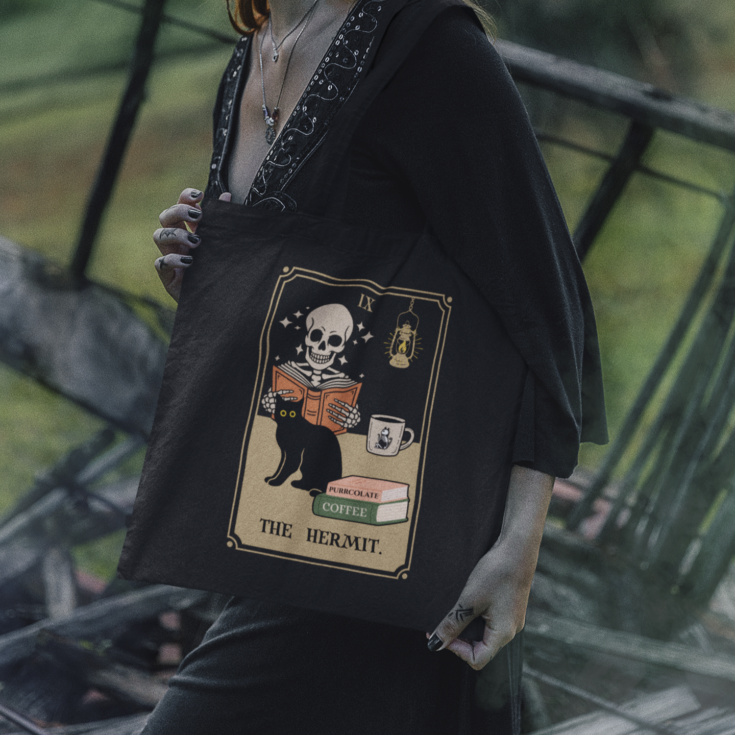 This tote features a parody of the tarot card The Hermit as a skeleton reading a book with a cat and a cup of coffee. In this image the tote is being carried on the shoulder of a woman who appears to be a witch.