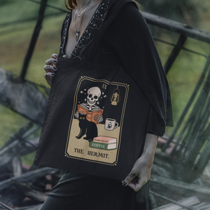 This tote features a parody of the tarot card The Hermit as a skeleton reading a book with a cat and a cup of coffee. In this image the tote is being carried on the shoulder of a woman who appears to be a witch.